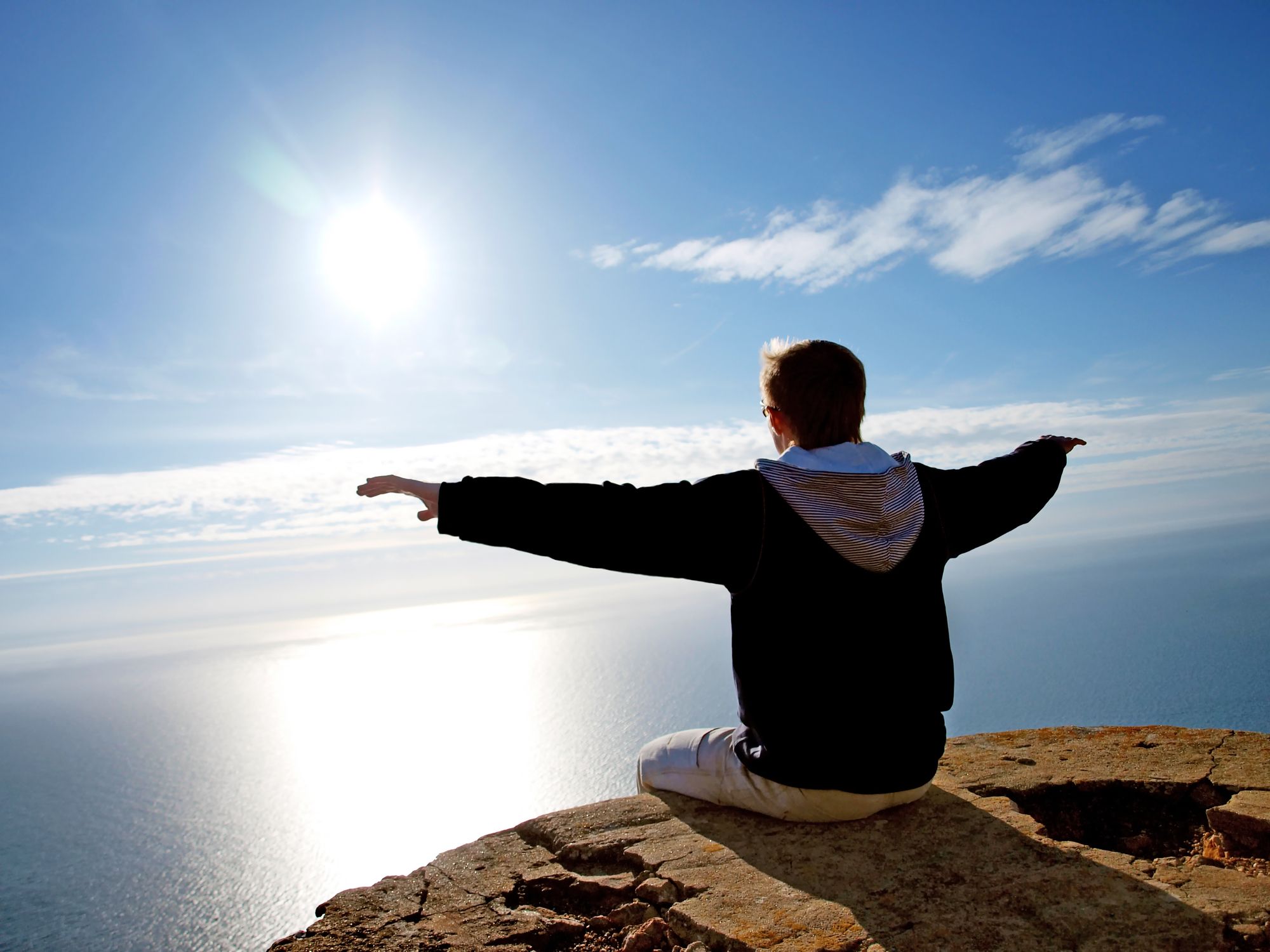 A man with his arms outstretched, sitting on a rock, facing the ocean and the sun.