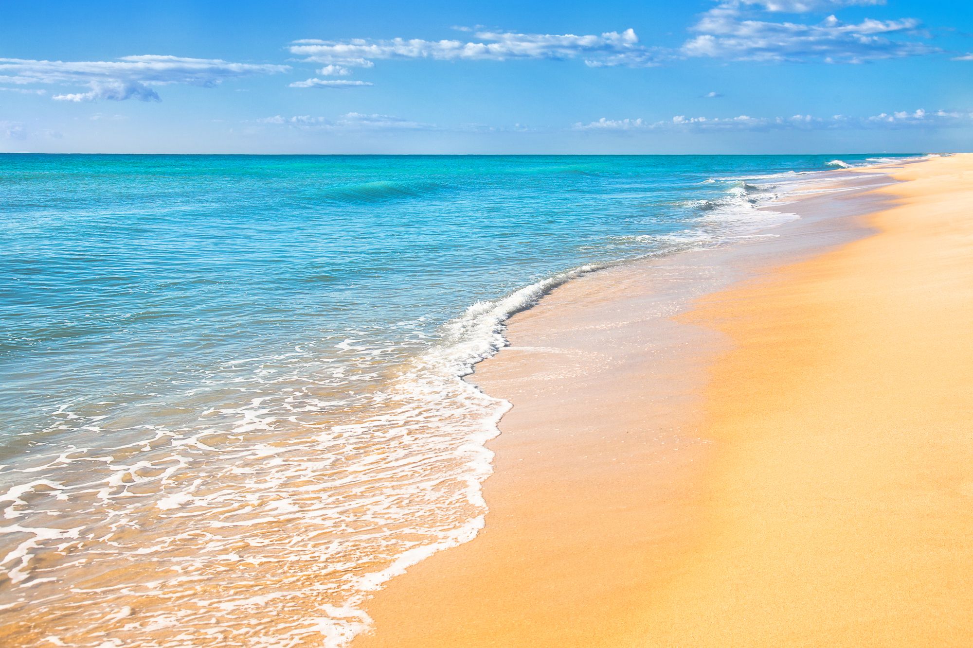 An idyllic beach scene, with golden sand, an aquamarine sea, and a bright blue sky with cumulus clouds.