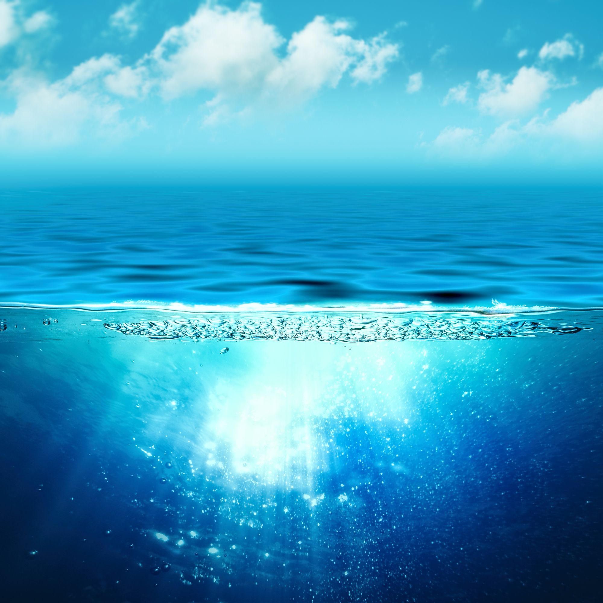 An ocean with a rippling blue surface and deep blue water below, where the area underwater is semi-penetrated by sunlight. The sky above is light blue with cumulus clouds.