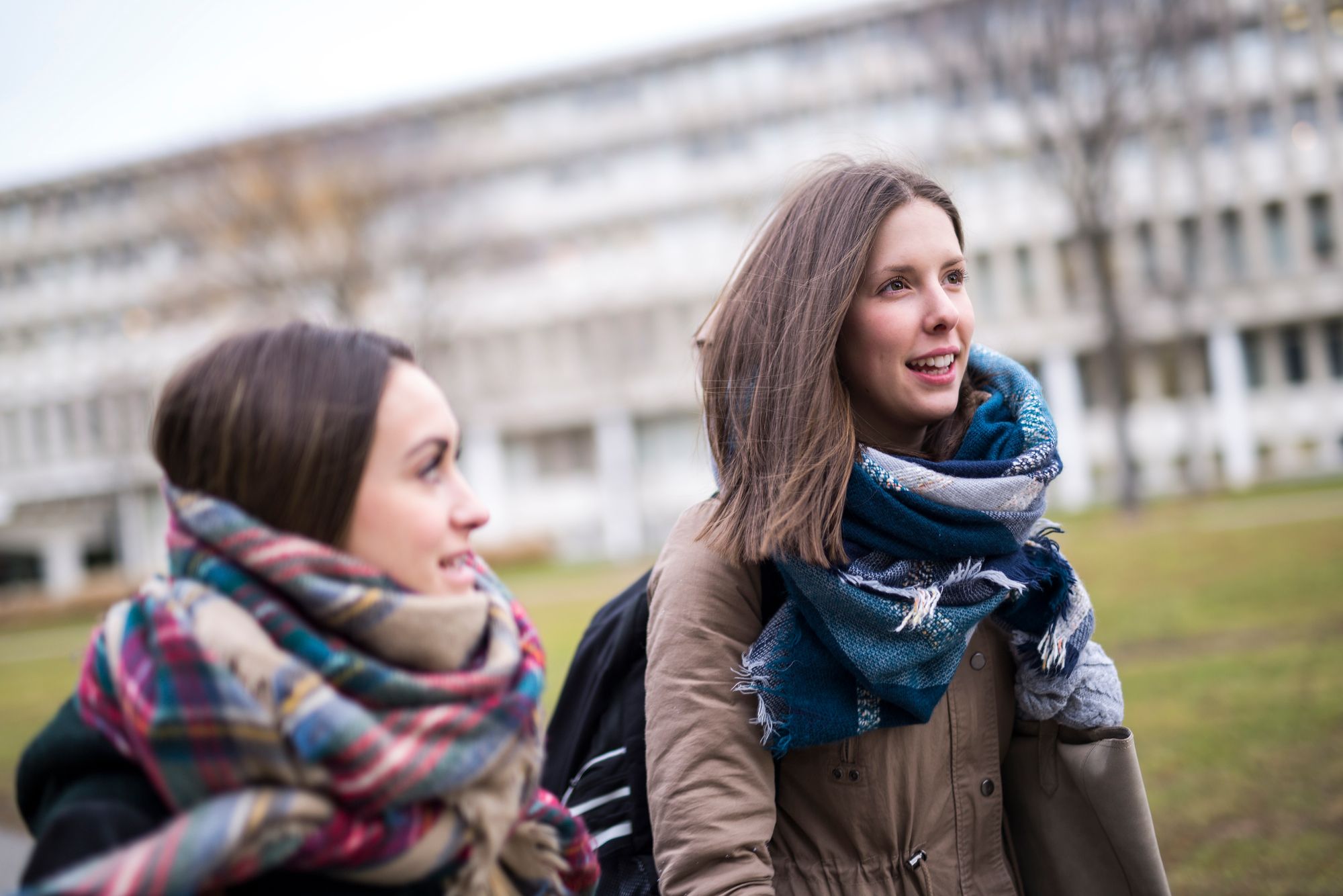 Two young women wearing jackets and scarves, walking out on campus, looking like a pair of happy friends.