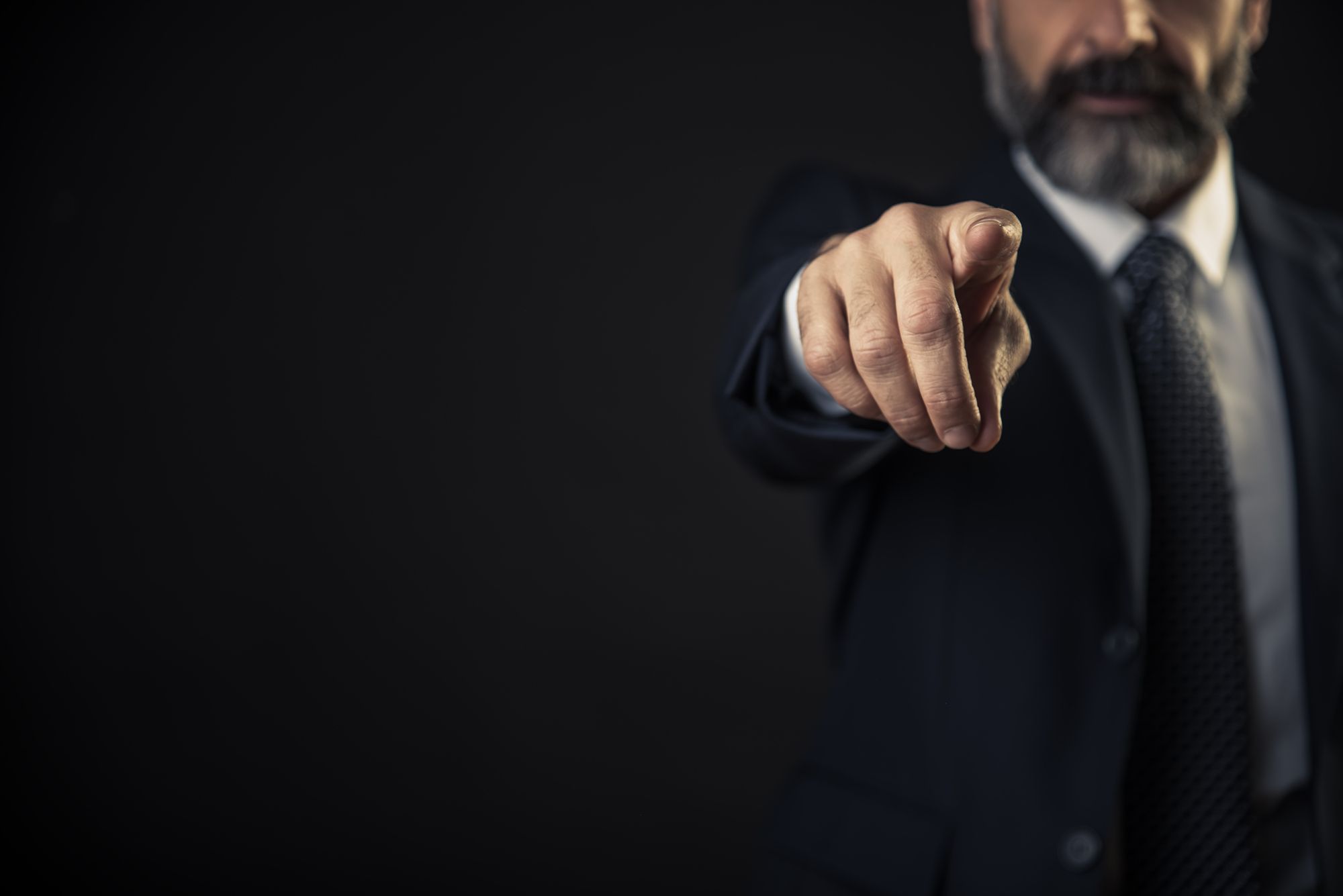 With a black background, a man in a suit and tie points a finger at the camera