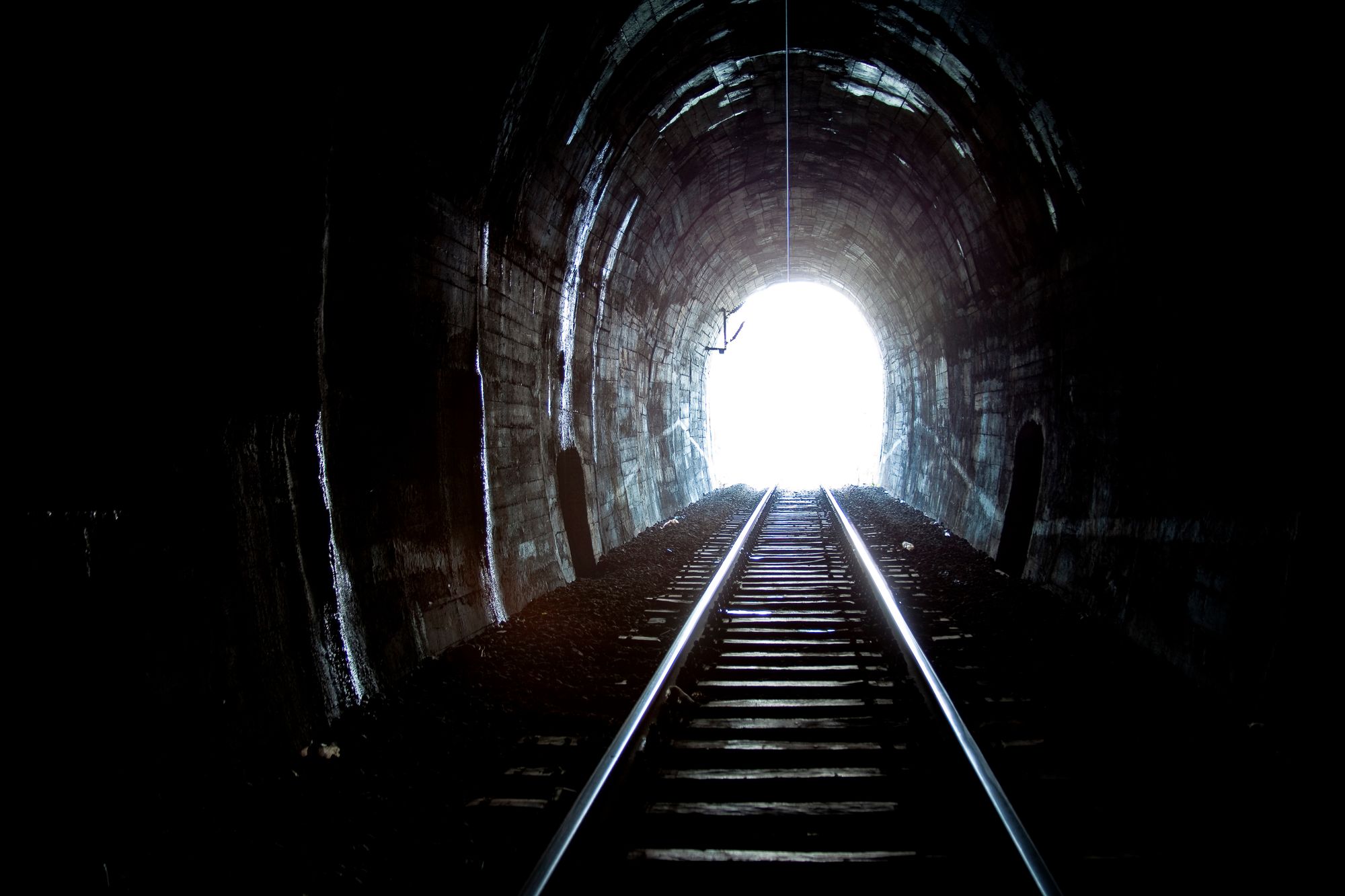 A dark tunnel with a train track, leading up to the light at the tunnel exit.