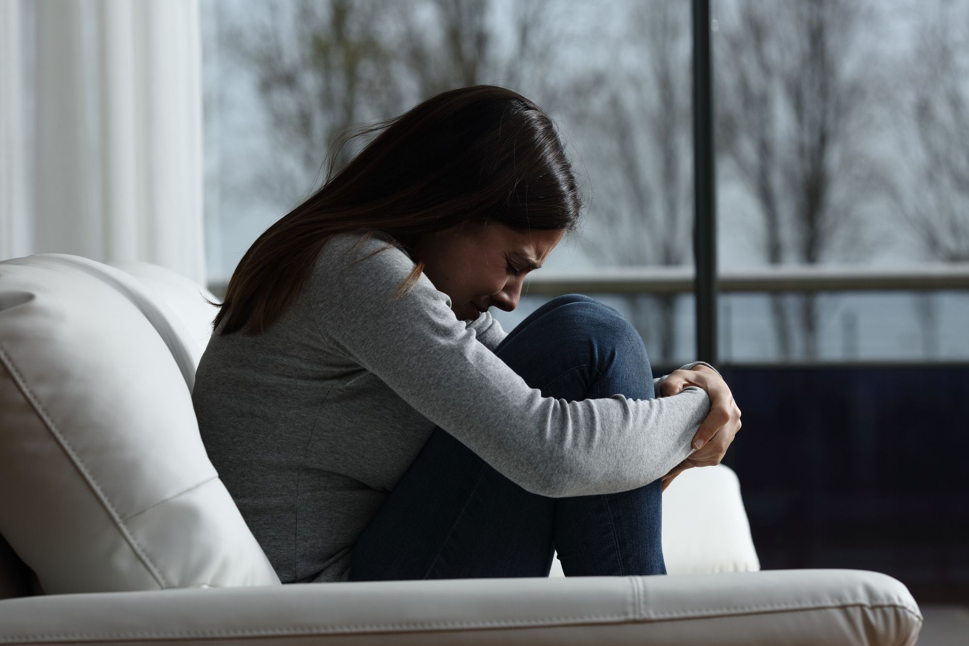 A girl with long black hair, is crying inconsolably, with her arms clutching her knees to her chest, sitting on a sofa.