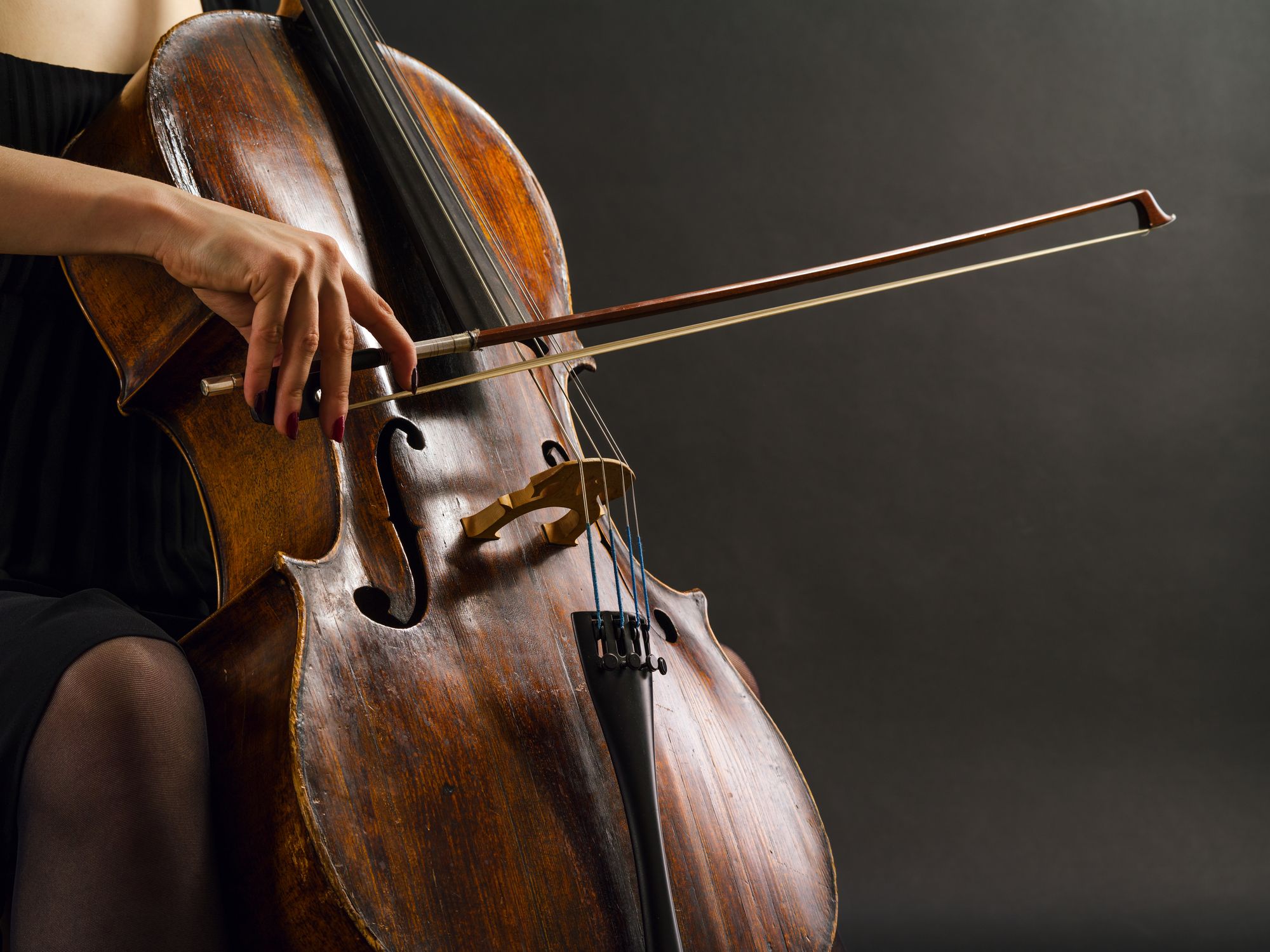 Someone plays a cello against a solid black background. Only the person's arm, neck, and thigh are visible.