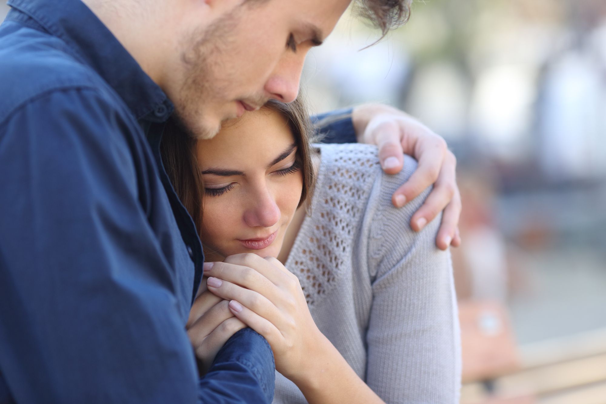 Man with navy blue shirt, hugs and comforts a sad woman in a grey shirt.