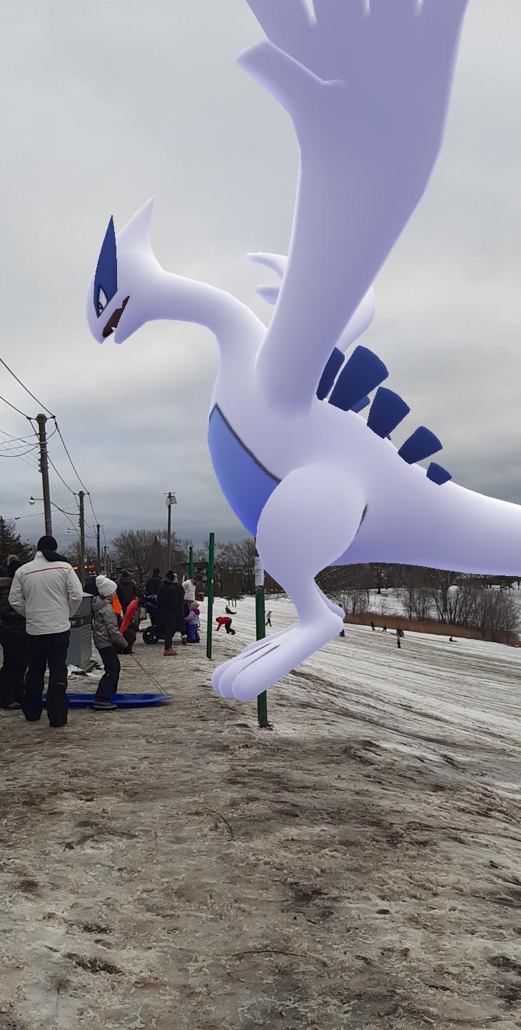 Lugia, the psychic and flying legendary bird Pokemon, flies above a hilly ground and a crowd of people.