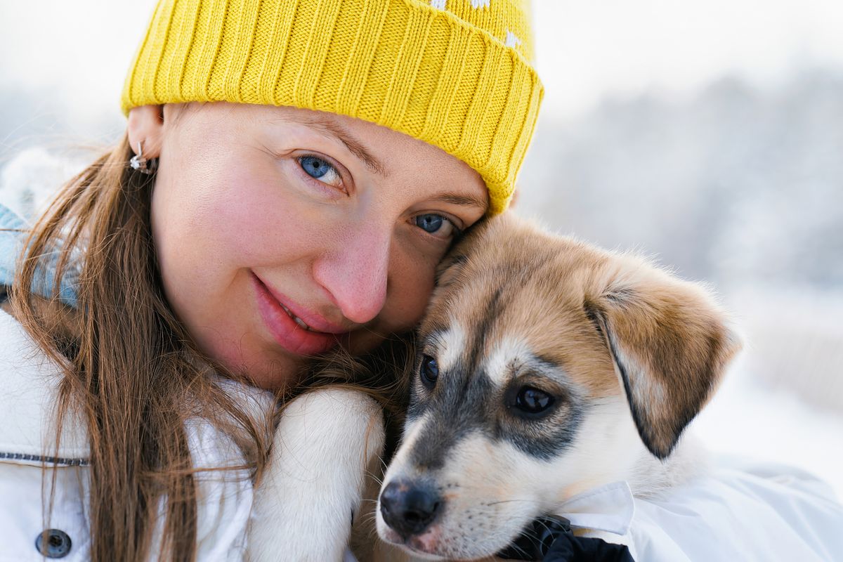 Woman with long brown hair and yellow beanie, white jacket, is smiling and hugging a husky puppy dog to her face. 