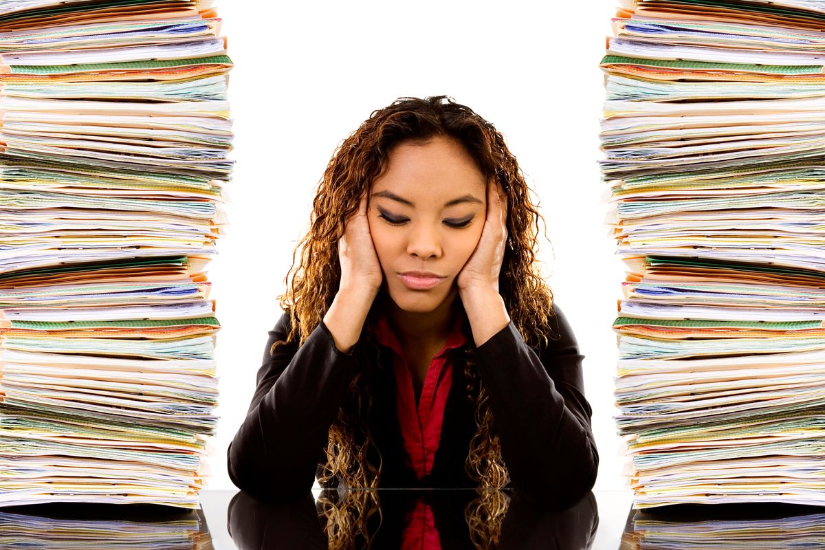 A woman holding her head in a headache, while stacks of papers pile up on either side of her.