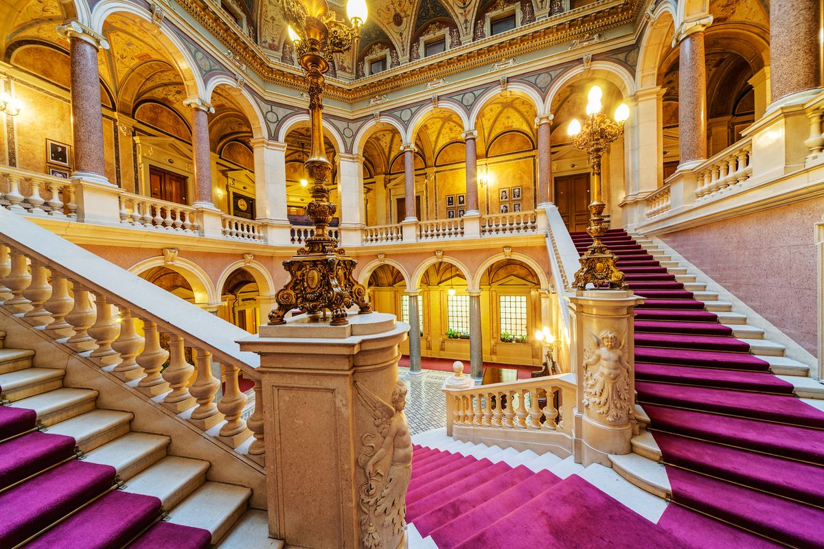 Interior of palace, full of golden, bright lights, and pinkish-violet carpets on the sweeping stairs.