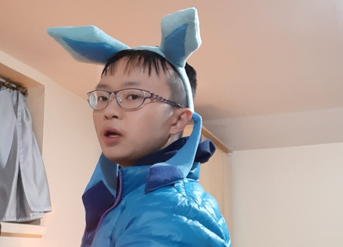 An Asian fellow wearing a blue puffy jacket, and a headband with two long blue ears and dangling blue ribbons.