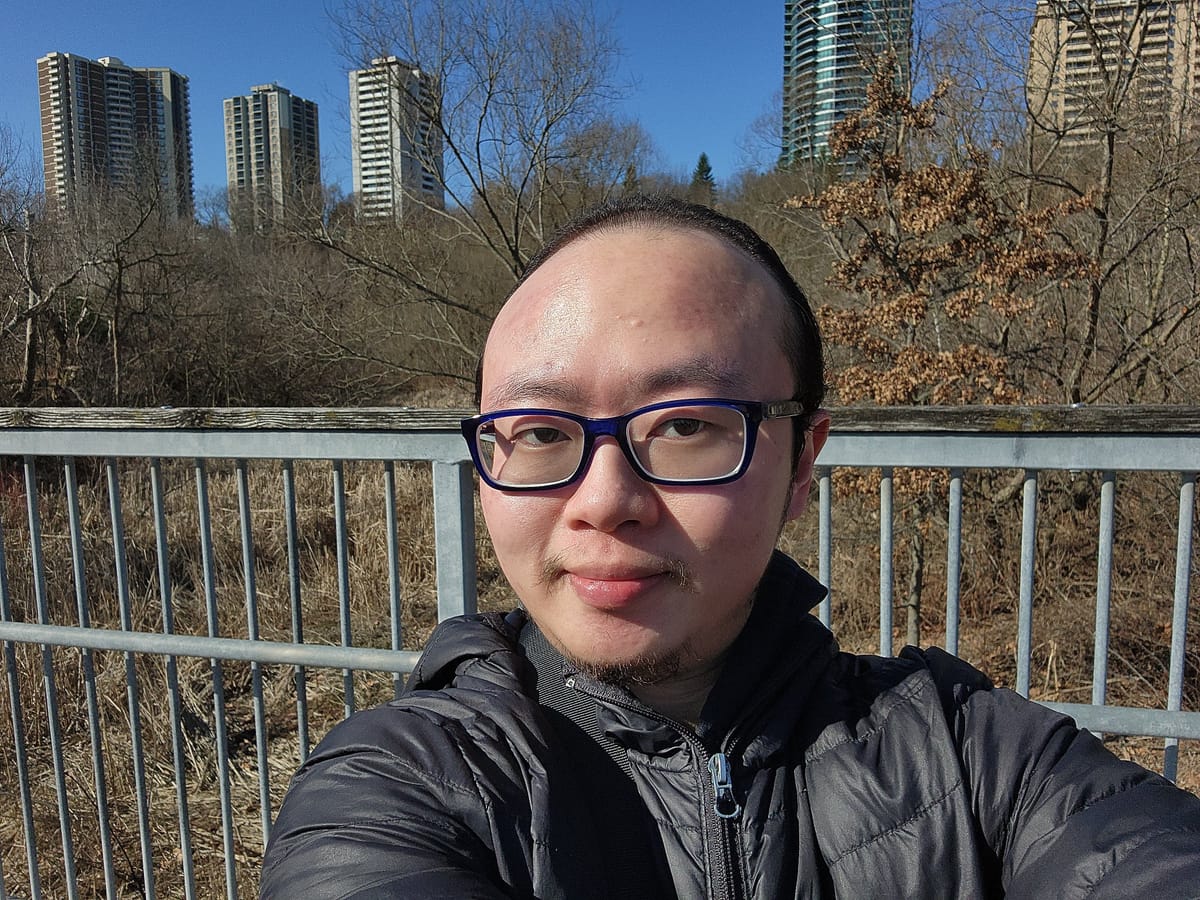 Asian man with glasses and black jacket, with a bridge in the background.