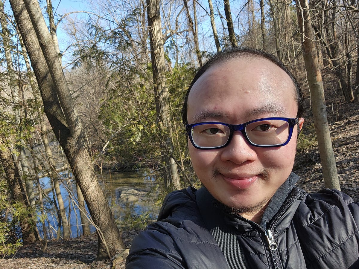 Asian man with glasses, smiling shyly, with trees and a brook in the background